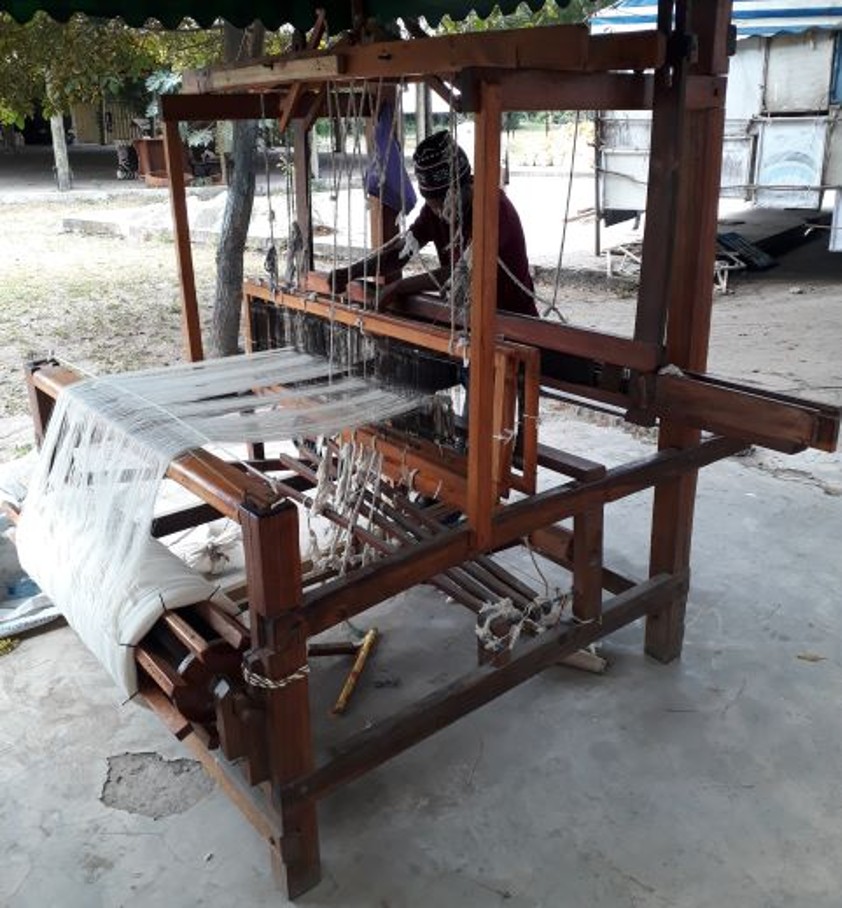 A handloom made of wood at which a man is processing yarn.