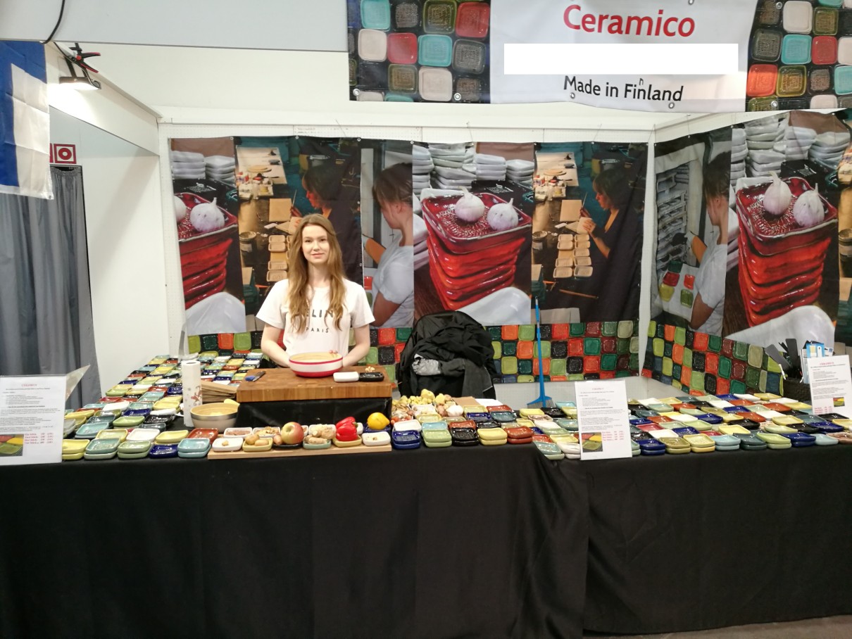 Eine Frau im T-Shirt an einem Messestand, davor zahlreiche Keramikreiben in quadratischer Form, im Hintergrund Poster vom Produktionsprozess. 