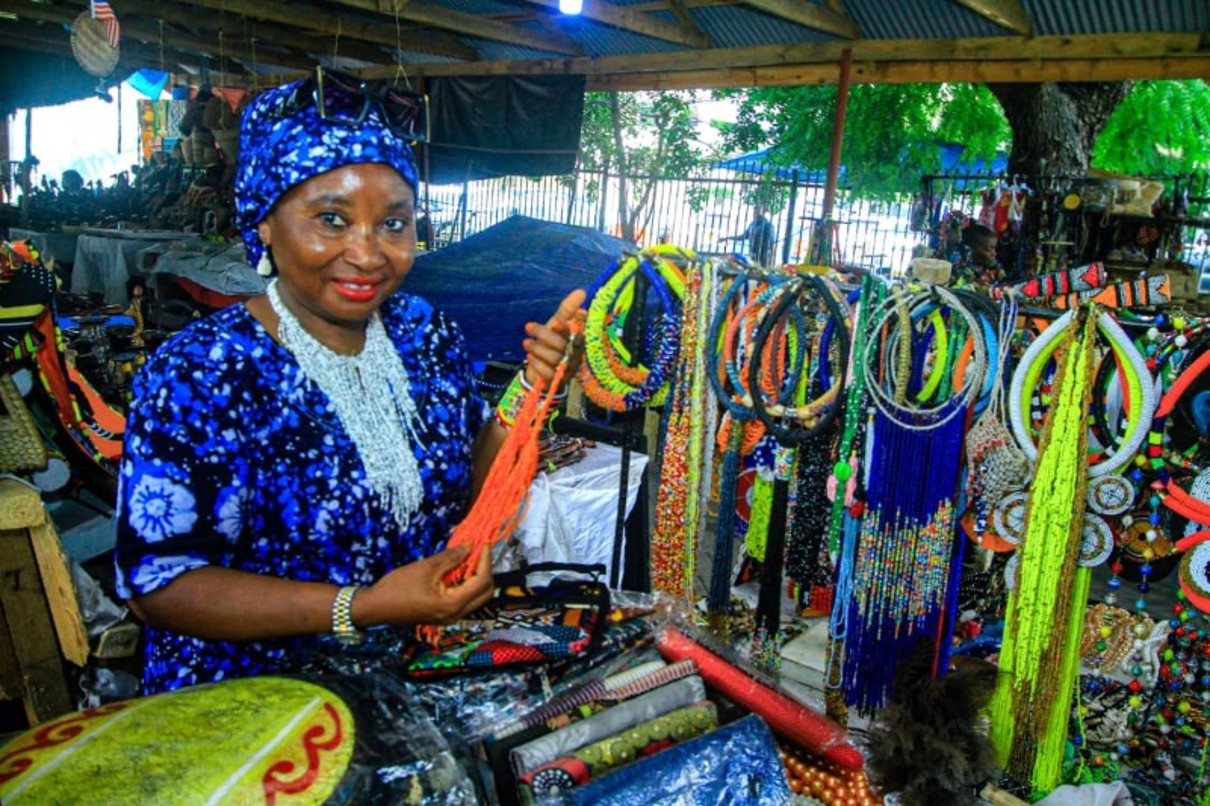 Eine Frau die lächelnd in die Kamera schaut und Perlenketten hochhält, umgeben von weiteren Produkten, wie Perlenketten und traditionellen Stoffen. 