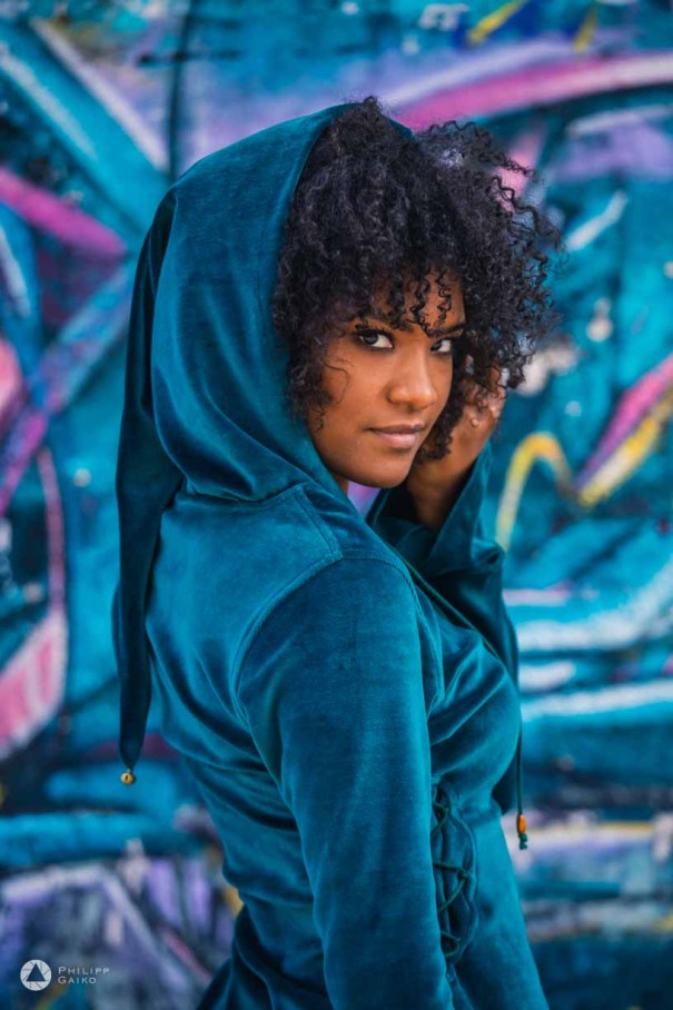 a woman in a blue jacket with hood standing in front of a colourful background.