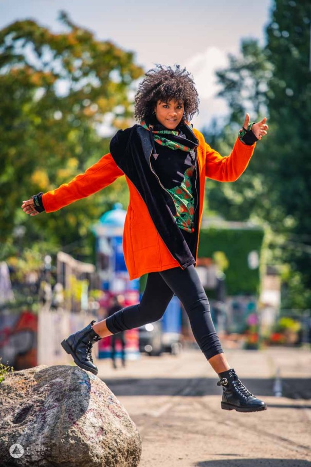 a woman in a red jacket jumps down from a stone and looks into the camera.