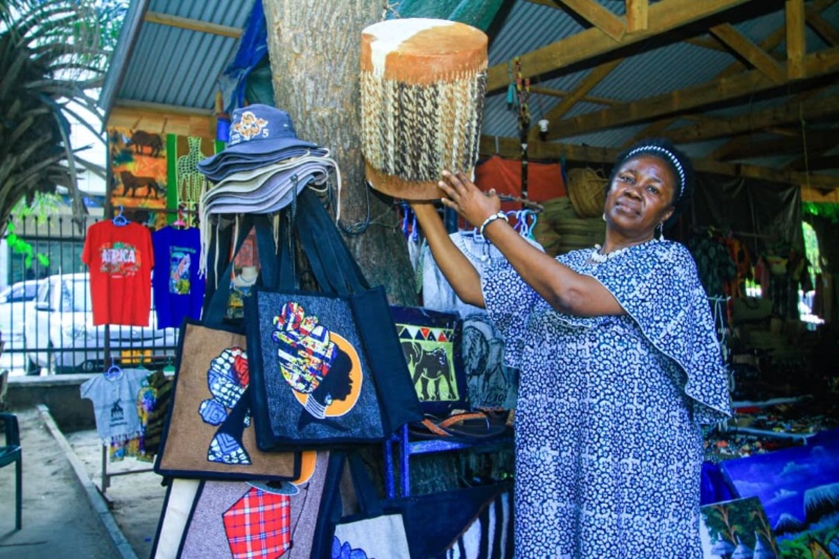 eine Frau, die eine handgearbeitete Trommel hochhält, neben ihr sind weitere Produkte zu erkennen, unter anderem Taschen mit traditionellen Mustern. 