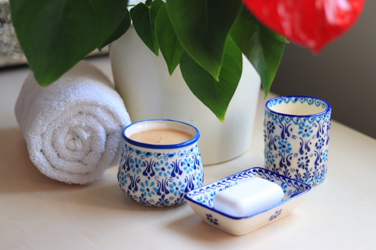 : a candle in a ceramic jar, a soap in a bowl and a ceramic mug in front of a flowerpot. Next to it lies a rolled-up towel. 