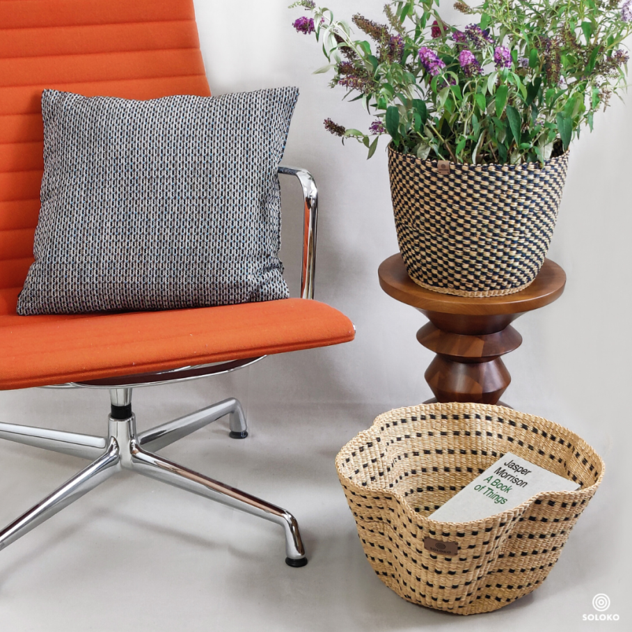 a chair with orange fabric on which a cushion is lying, next to it a table with a plant basket, another basket is on the floor. 