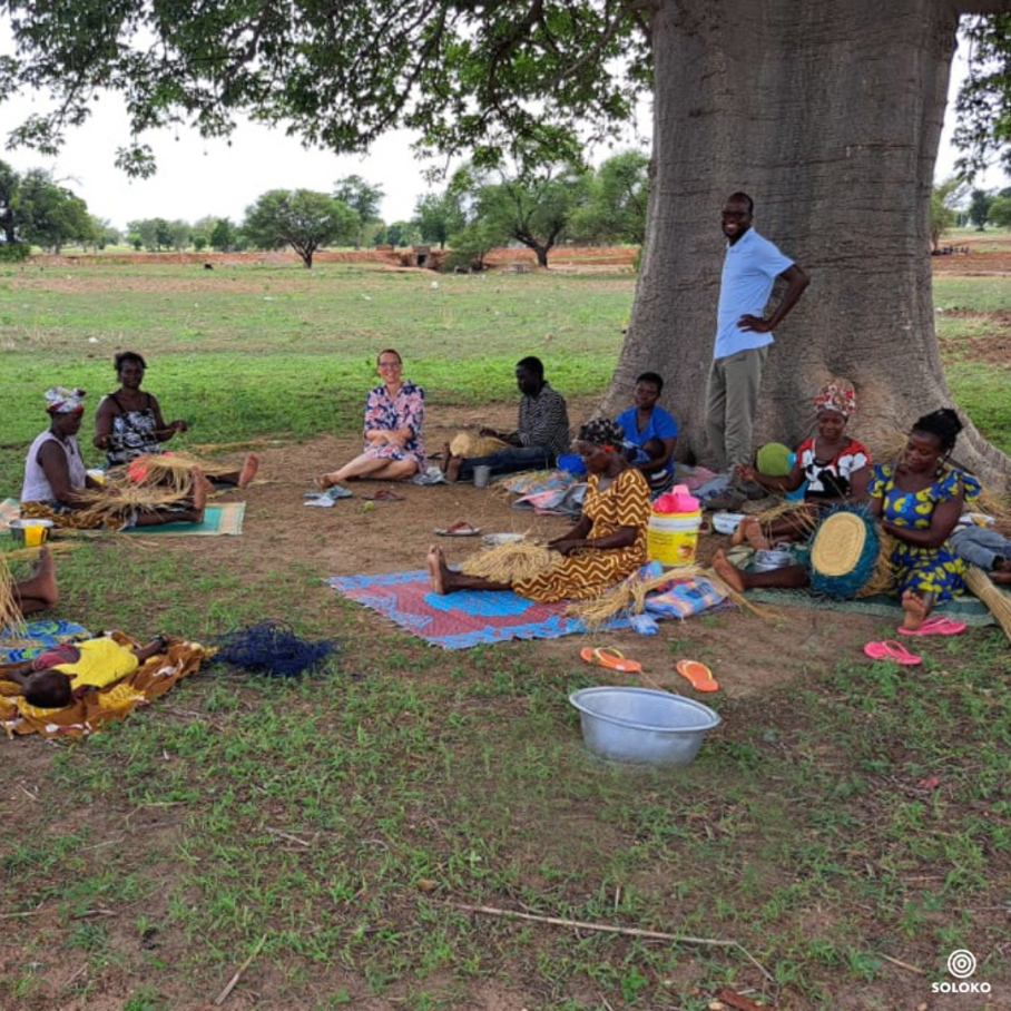 A group of people sits on grass in front of a tree and weaves baskets. 
